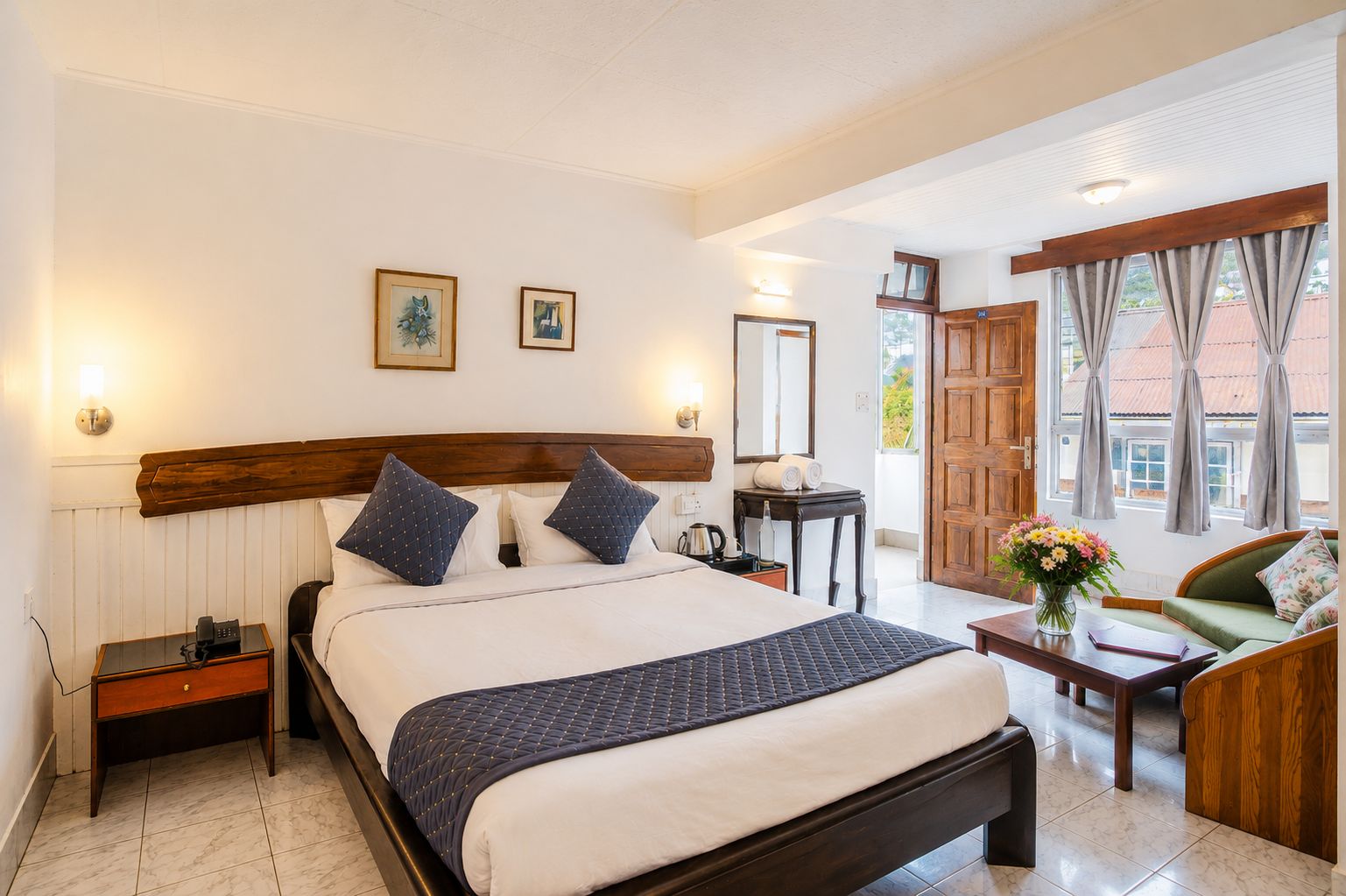 Habitación de hotel con una cama grande, mesitas de noche, zona de estar con sofá y sillón, una pequeña mesa con un jarrón de flores, y vistas al exterior a través de una ventana.