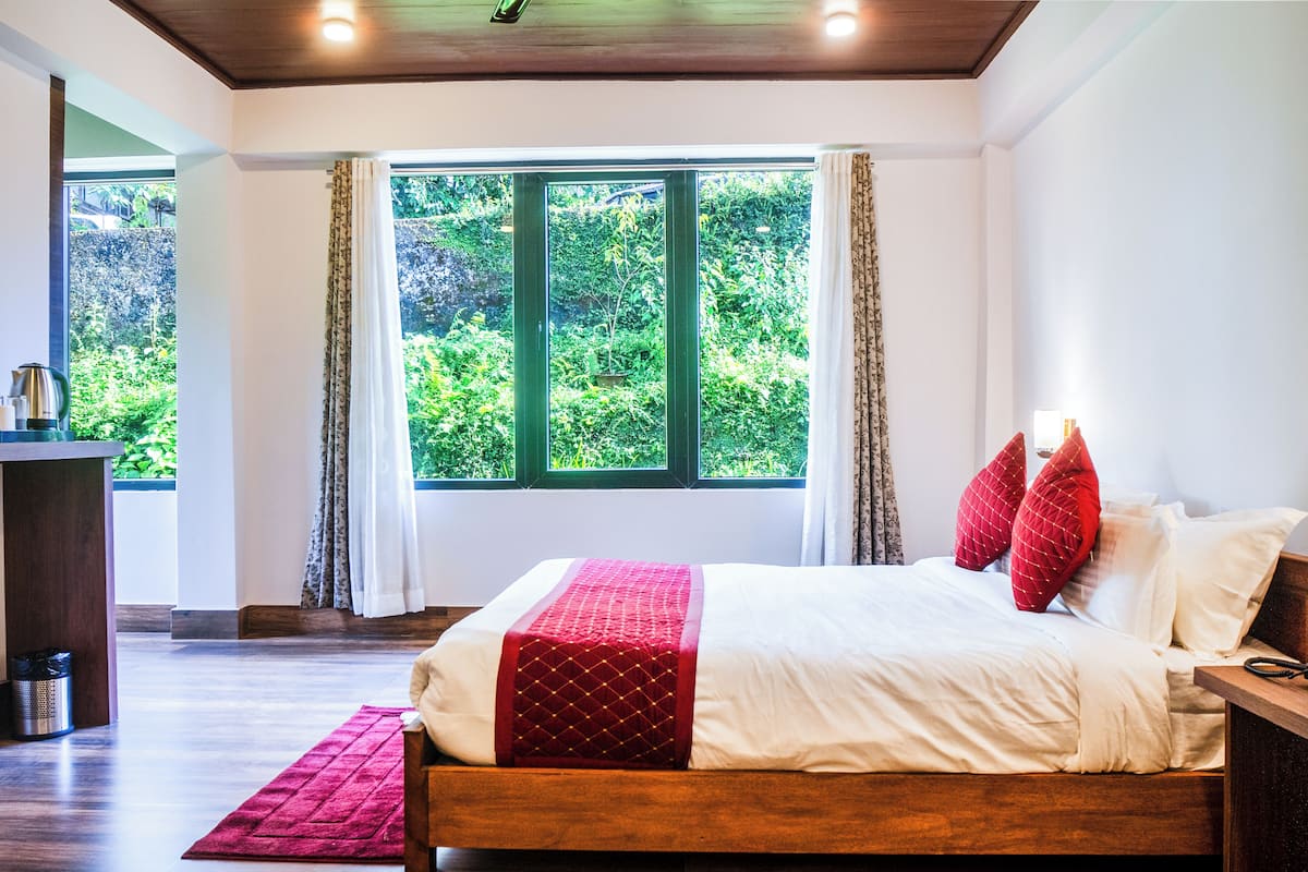 A bedroom with a wooden bed, a red and white bedspread, and a view of lush greenery through a large window.