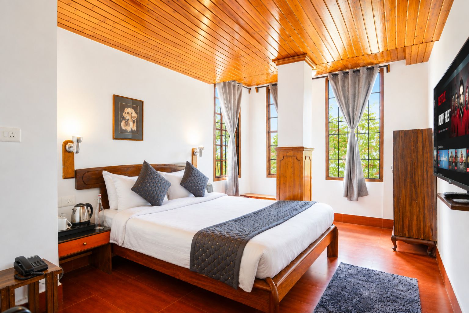 A hotel room with a wooden ceiling, a bed with a quilted blanket, a wooden nightstand with a telephone, a wooden wardrobe, and a television on the wall.
