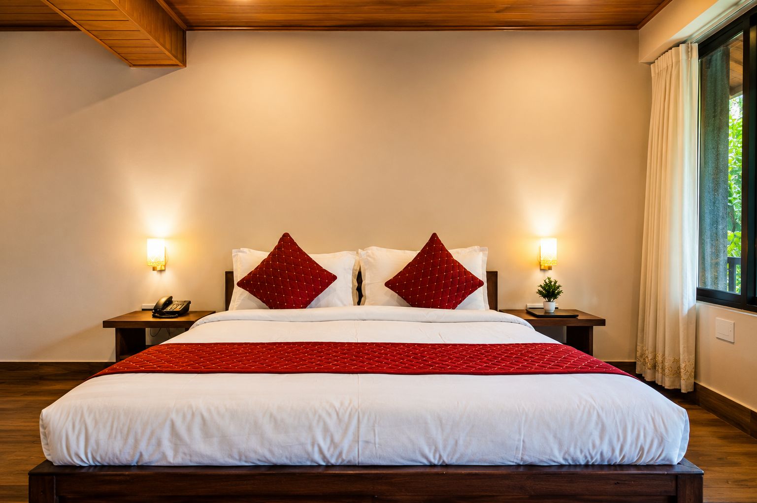 Une chambre d’hôtel avec un grand lit, deux tables de chevet, un téléphone, une plante et une fenêtre avec des rideaux.