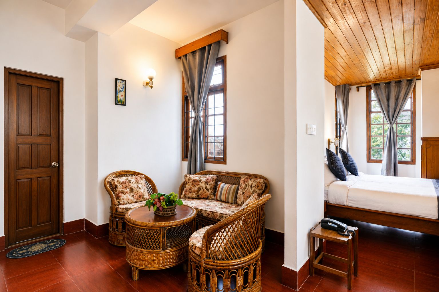 A room with a wooden door, a sofa with floral cushions, a small table with a plant, and a bed with a wooden headboard.