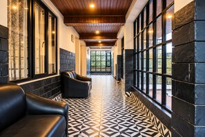 A hallway with black leather sofas, patterned floor tiles, and large windows with a view of greenery.