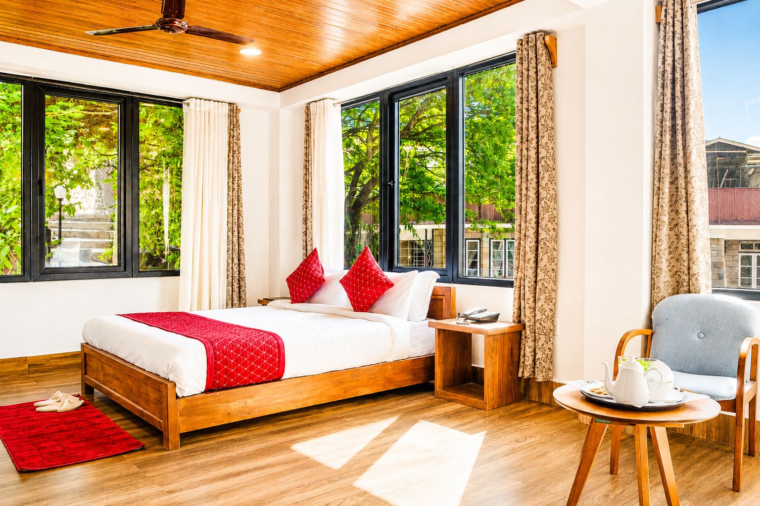 A hotel room with a wooden bed, a bedside table with a phone, a chair, and a view of greenery outside.