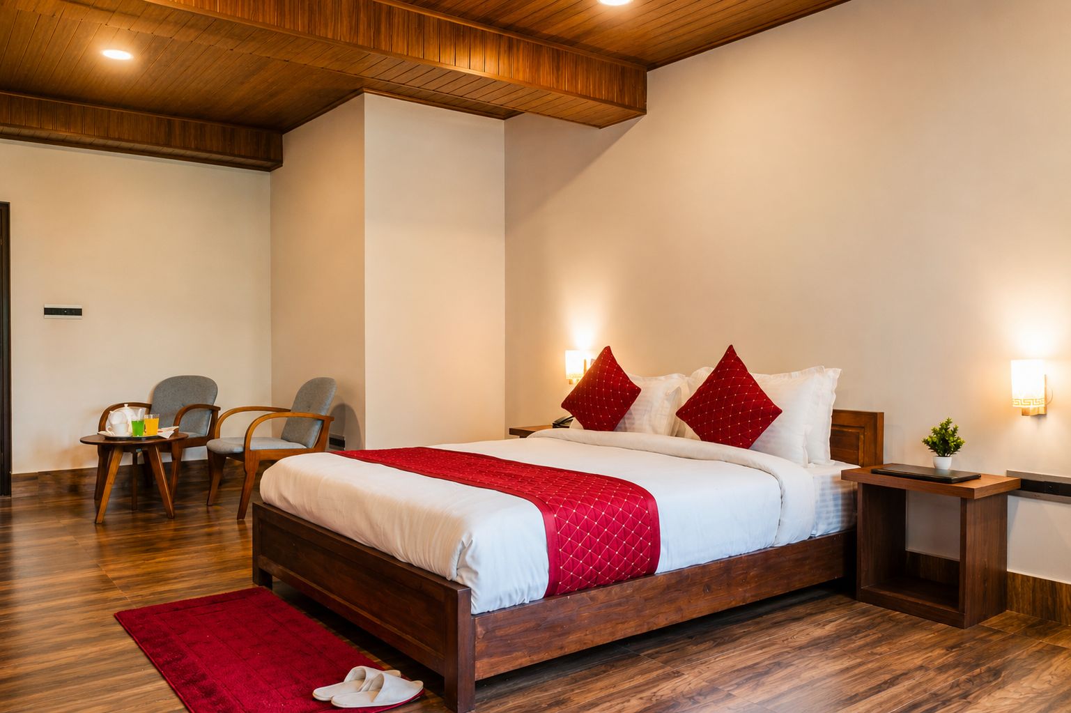 A hotel room with a wooden bed, a red throw blanket, a nightstand, a chair, and a small table with a plant.