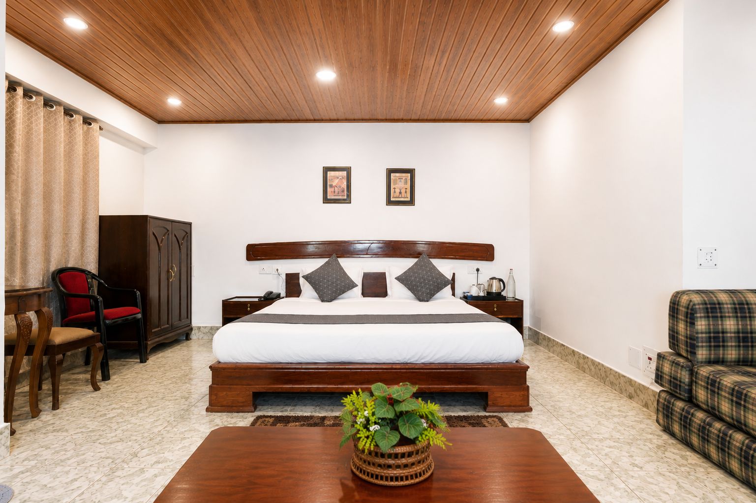 Ein Hotelzimmer mit einem großen Bett, einem Holz-Kopfteil, zwei Nachttischen, einem roten Sessel, einem Kleiderschrank, einem karierten Sofa und einem Holztisch mit einer Pflanze.