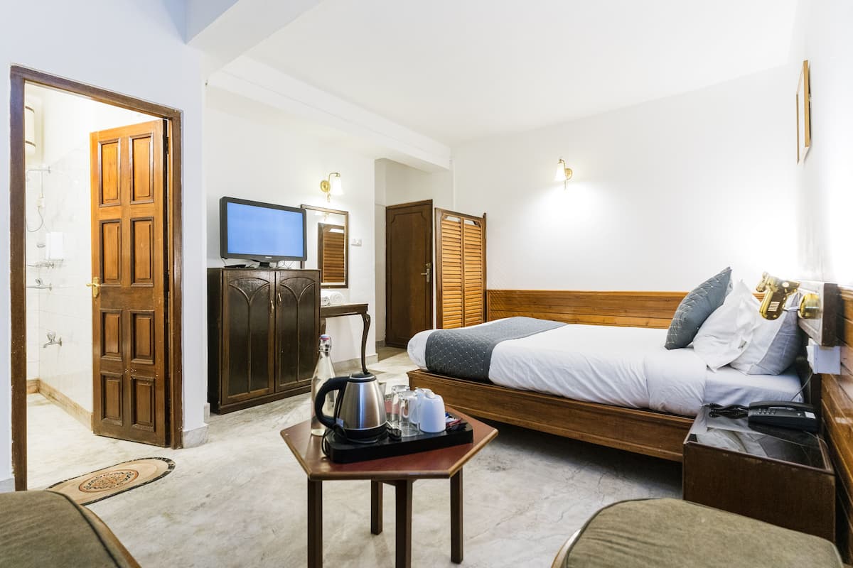A hotel room with a bed, a television, a wooden wardrobe, a small table with a kettle, and a bedside table with a phone.
