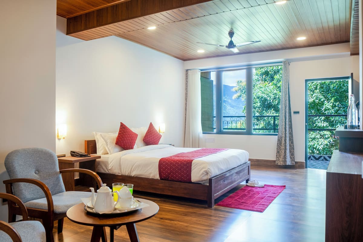 A hotel room with a wooden ceiling, a large bed with red pillows, a bedside table with a phone and a glass of juice, and a view of greenery through the window.