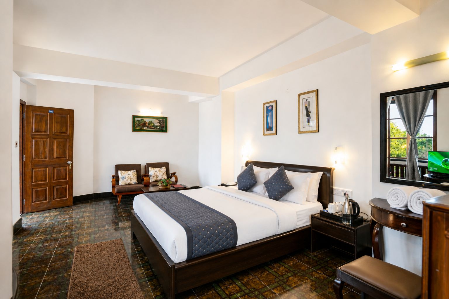 A hotel room with a large bed, a sitting area with chairs, a desk with a computer, and a window with a view.