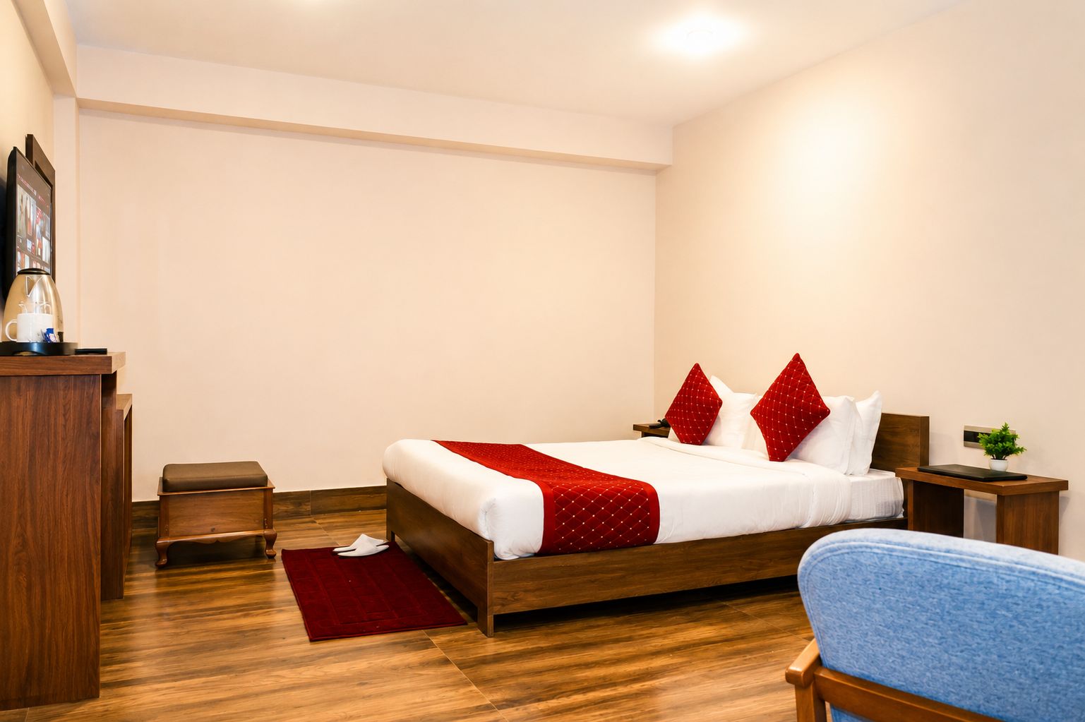 Una camera d'albergo con un letto di legno, una coperta rossa, un comodino in legno, un tavolino e una sedia blu.