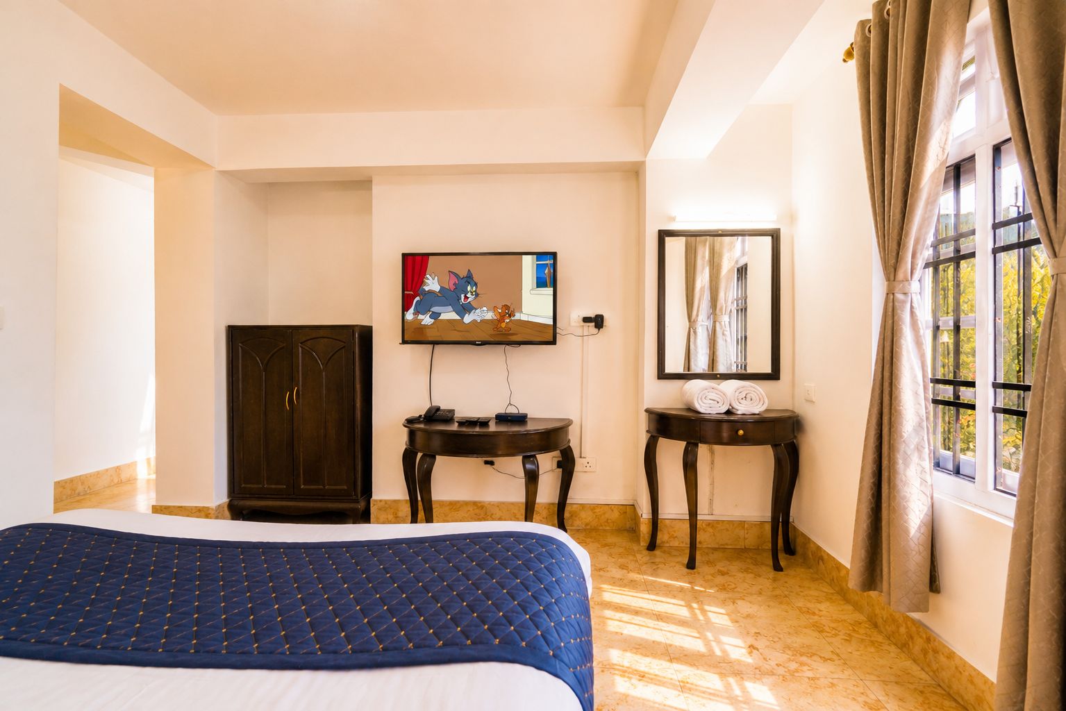 Habitación de hotel con cama, televisor, escritorio, armario y ventana con cortinas.
