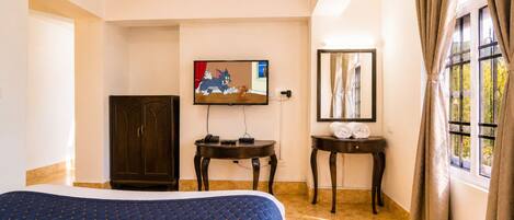 Quarto de hotel com cama, televisão, escrivaninha, guarda-roupa e janela com cortinas.