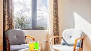 Una habitación con dos sillas, una mesa con una bandeja de galletas y una tetera, y una ventana que da a vistas de árboles y nubes.