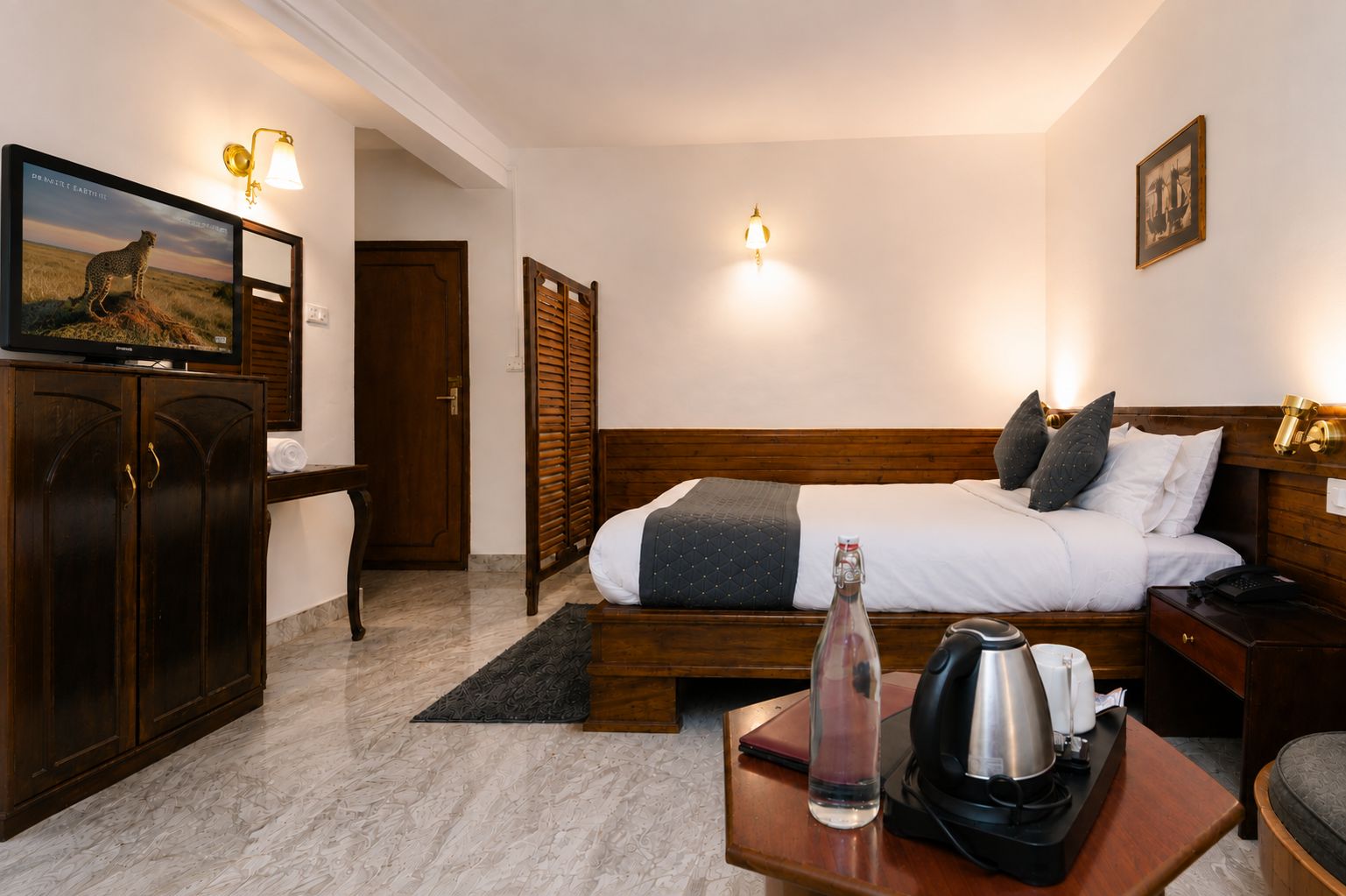 A hotel room with a bed, a desk with a kettle, a bottle, and a picture on the wall.