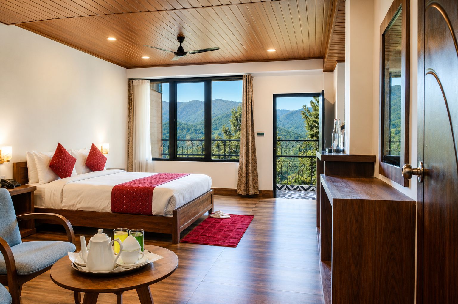 Una habitación de hotel con una cama, un escritorio, una silla, una mesa con tetera y tazas, y ventanas amplias con vista a la montaña.
