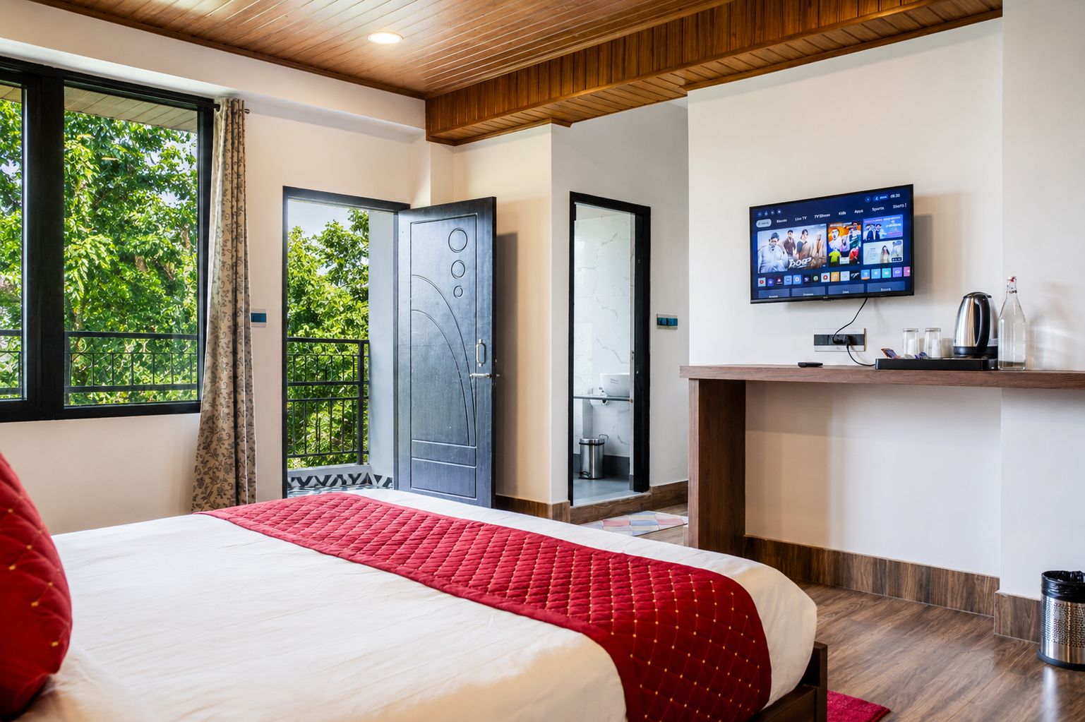 A hotel room with a bed, a TV mounted on the wall, a door, and a window with a view of greenery.