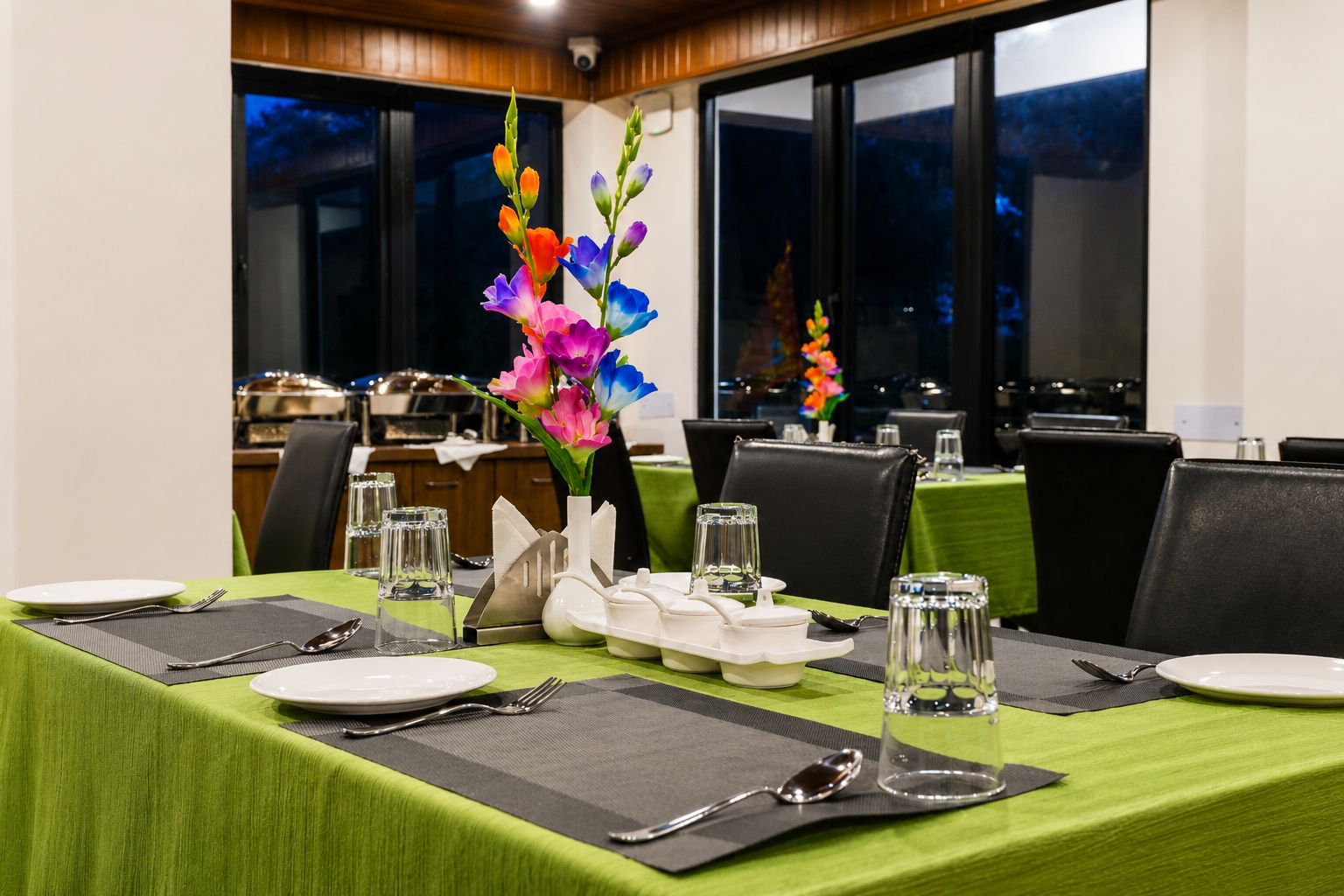 A dining table set with green tablecloths, black chairs, and a vase of colorful flowers.