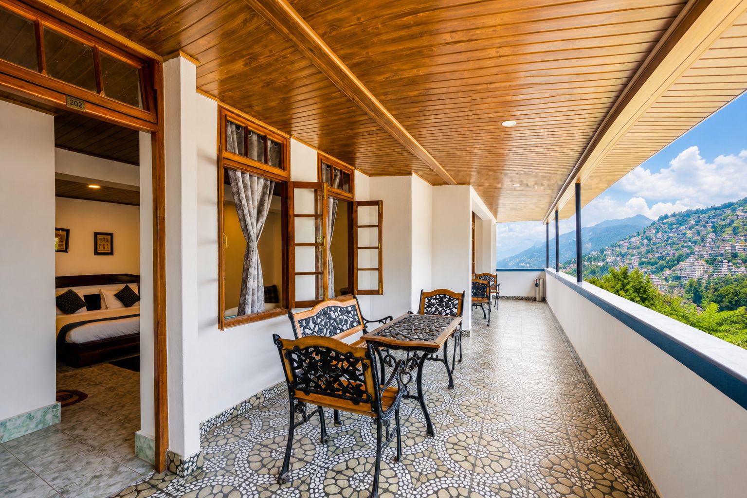 A balcony with a tiled floor, wooden ceiling, and a bed with a headboard visible through an open window.