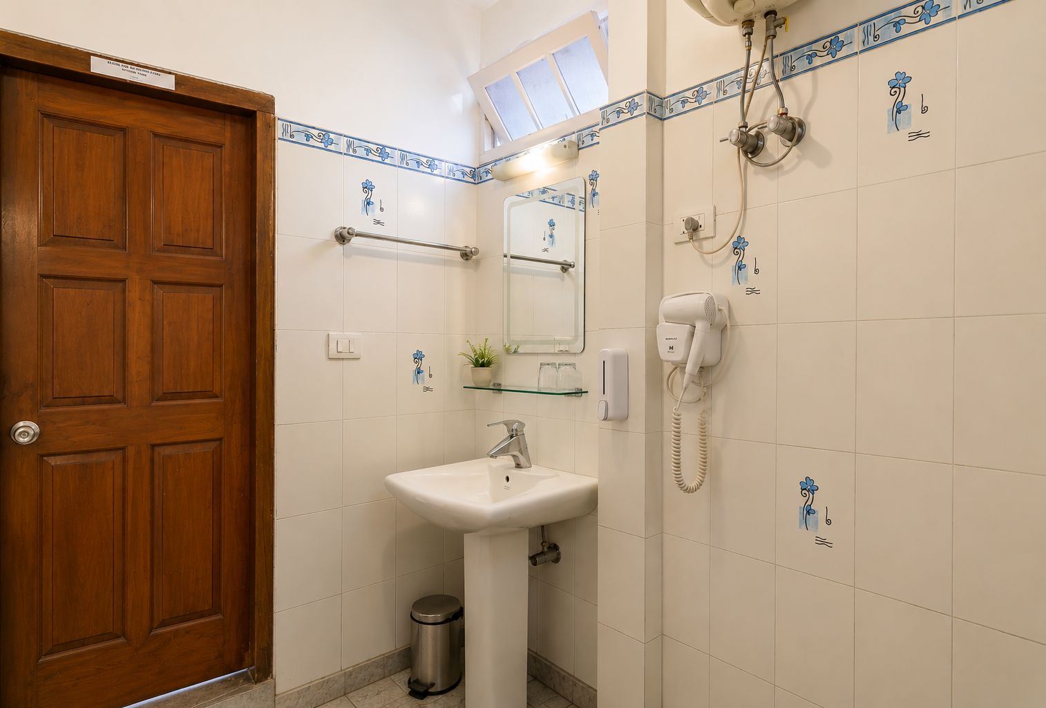 A bathroom with a wooden door, a white sink, a mirror, a towel rack, a shower, and a trash bin.