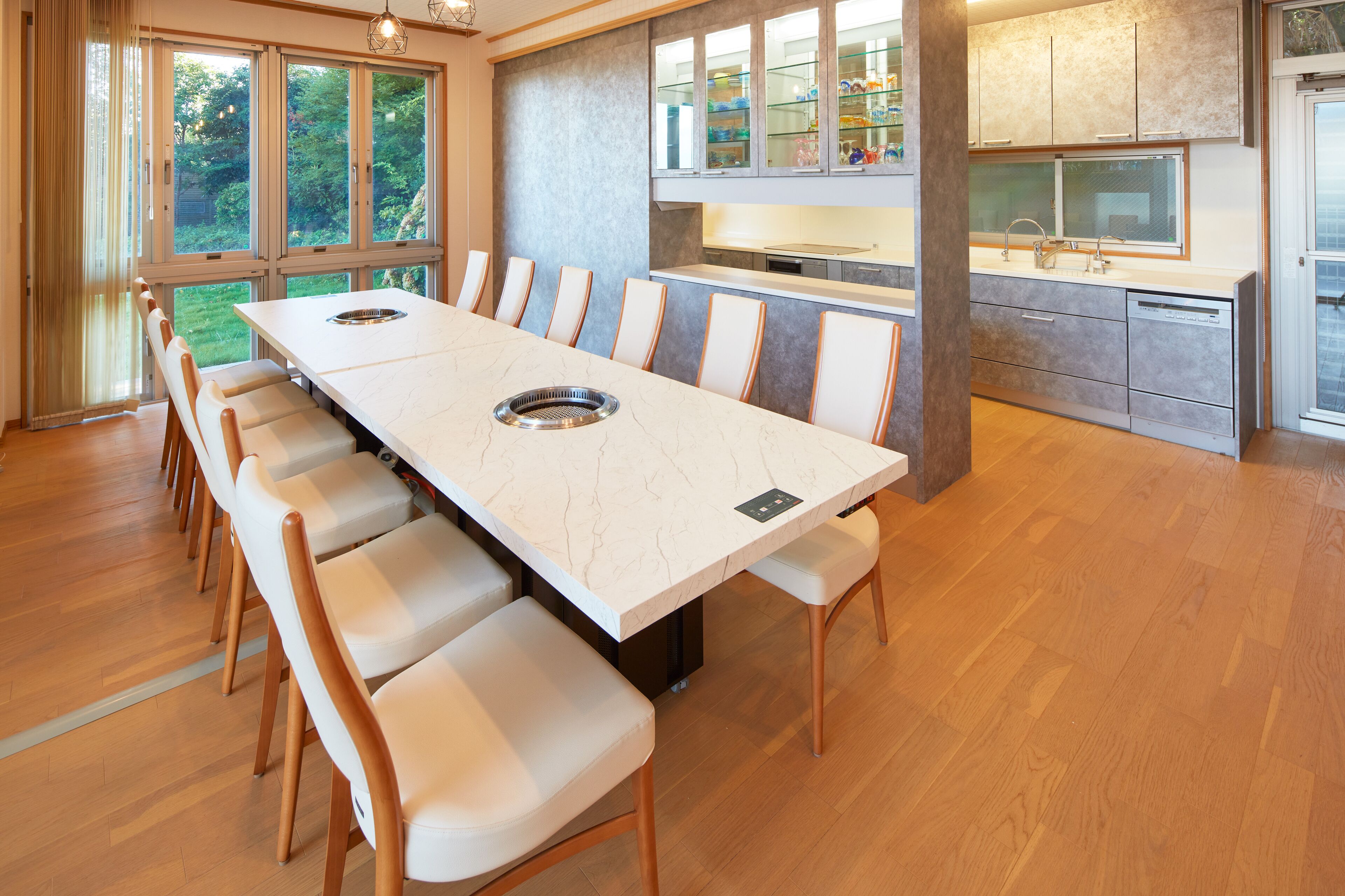 A modern kitchen with a large island, a dining area with a long table and chairs, and a view of greenery outside.