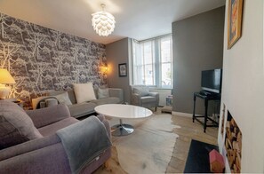 A living room with a sofa, a coffee table, a lamp, a television, and a patterned wallpaper. - York House is a three-bedroom house with parking and convenient access to both town and beach. (Whitstable)