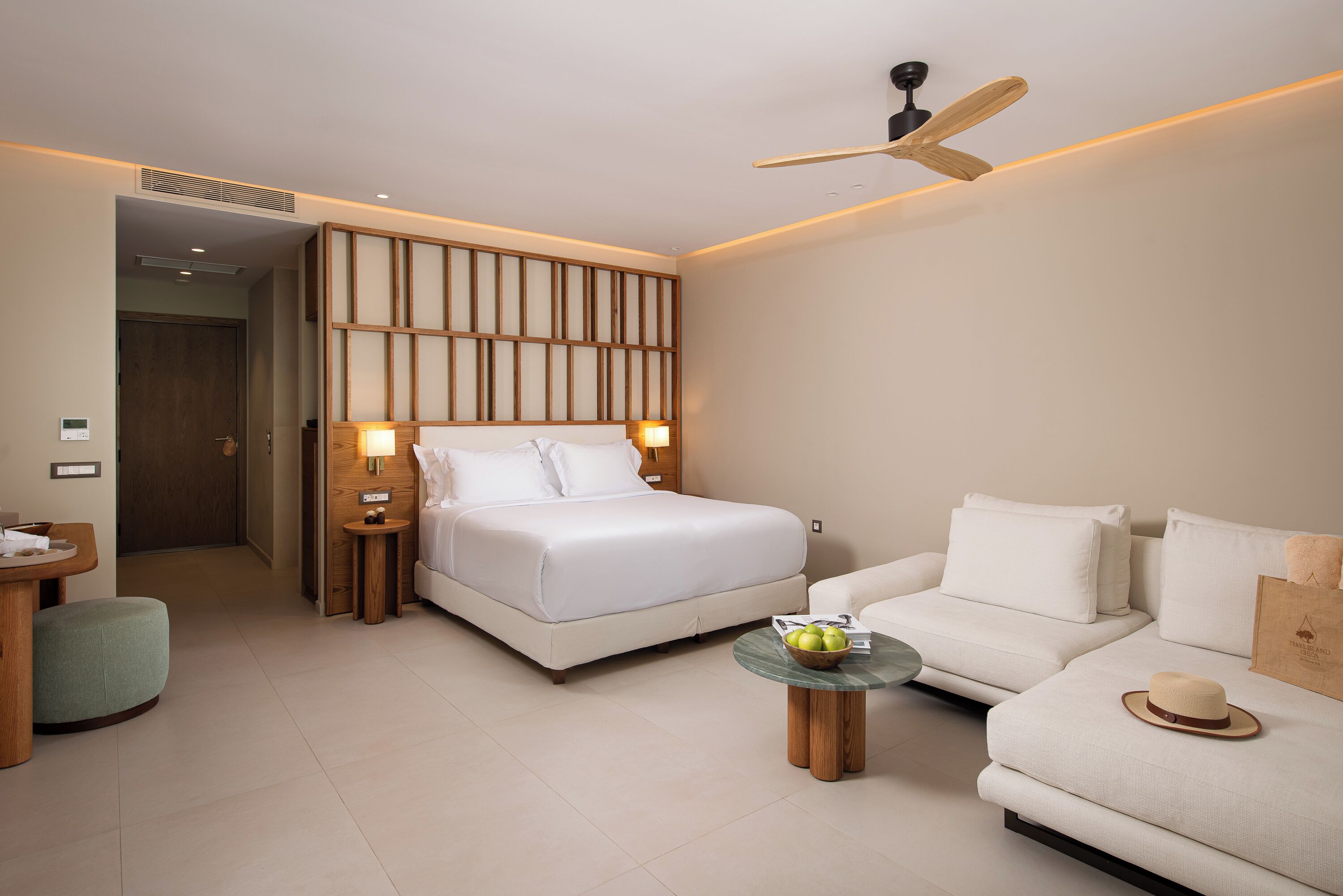 A modern hotel room with a large bed, a sofa, a small table with a bowl of fruit, and a ceiling fan.