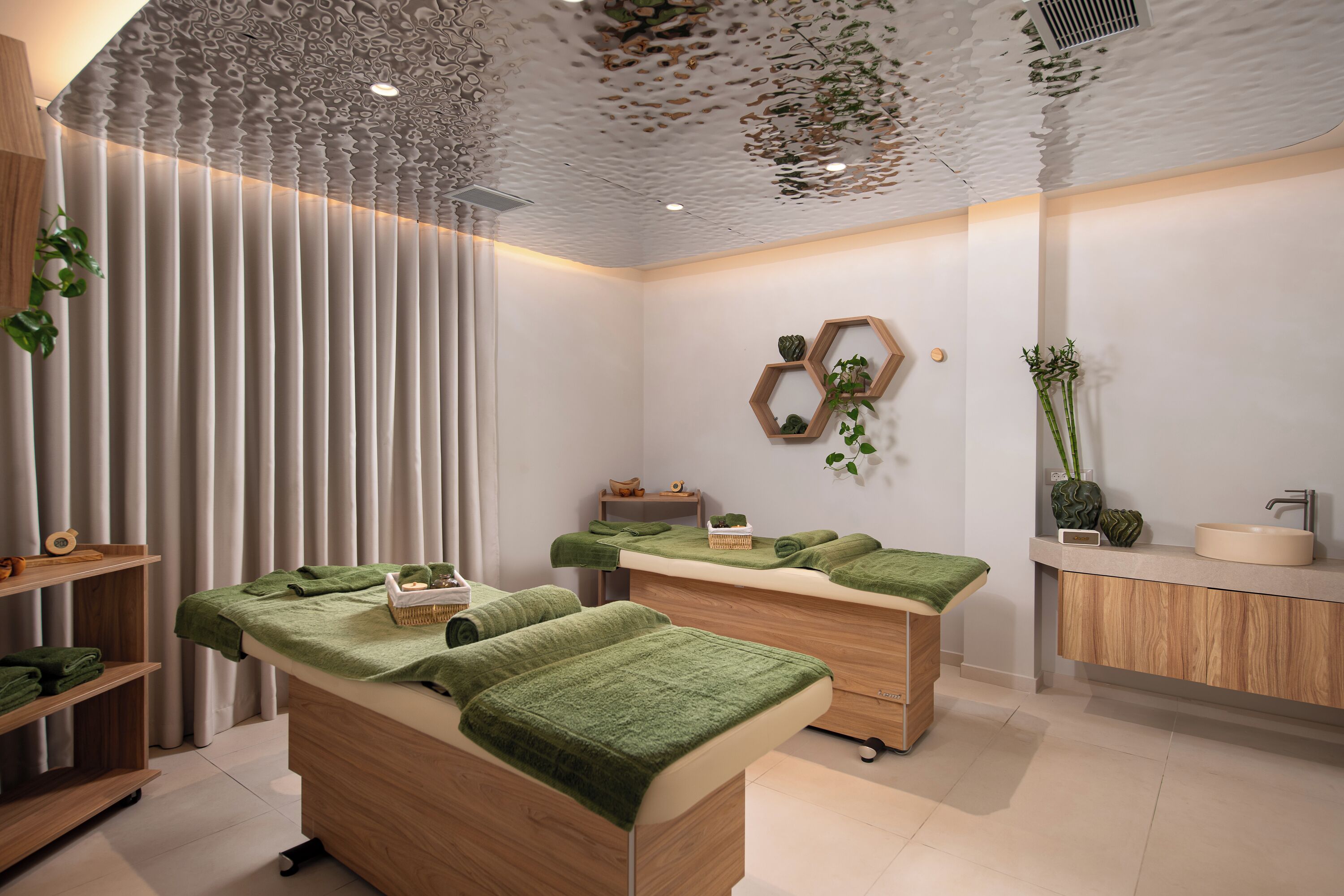 A modern spa room with two massage tables, a wooden cabinet, and a wall-mounted hexagonal shelf with plants.