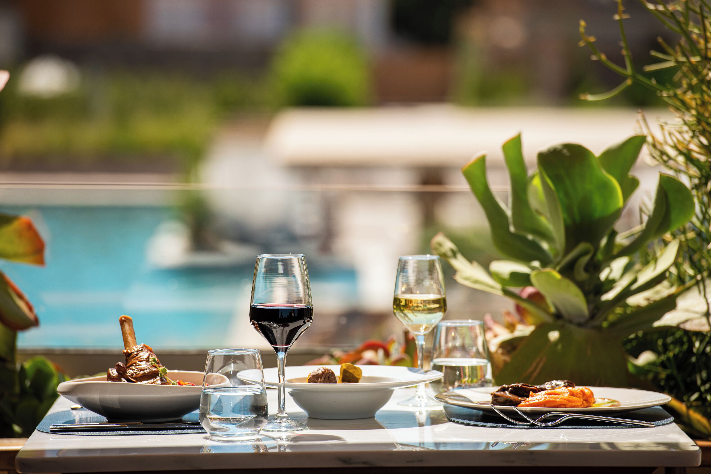 Une table à manger avec des assiettes remplies de nourriture, des verres de vin et une piscine en arrière-plan.