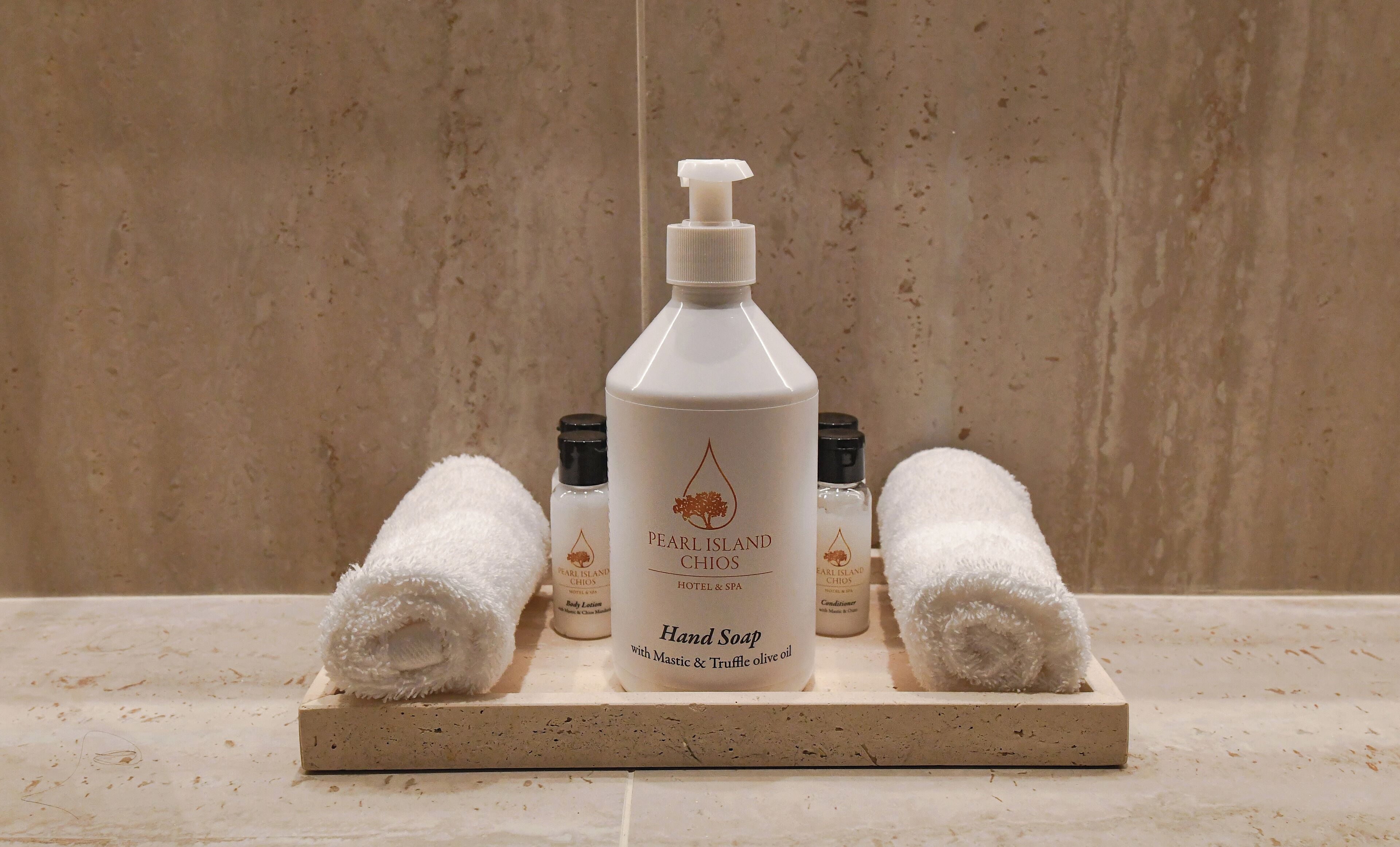 A bottle of hand soap with a pump dispenser, two rolled towels, and several smaller bottles of body lotion on a wooden tray.