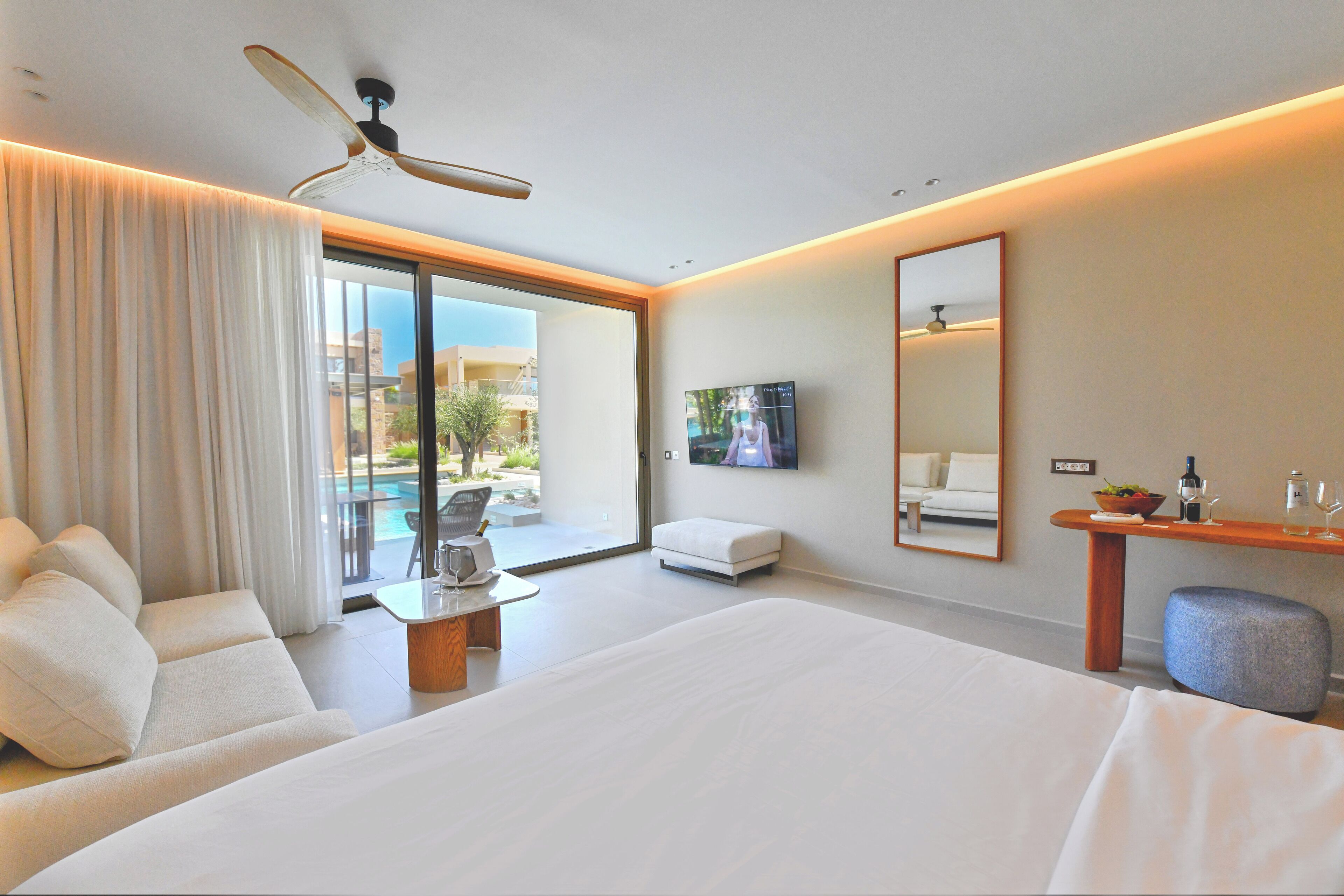 A modern hotel room with a large bed, a sofa, a TV, a ceiling fan, and a view of the outdoors.