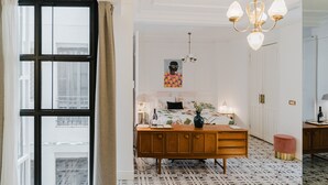 A bedroom with a large window, a wooden dresser, a bed with floral bedding, a chandelier, and patterned flooring.