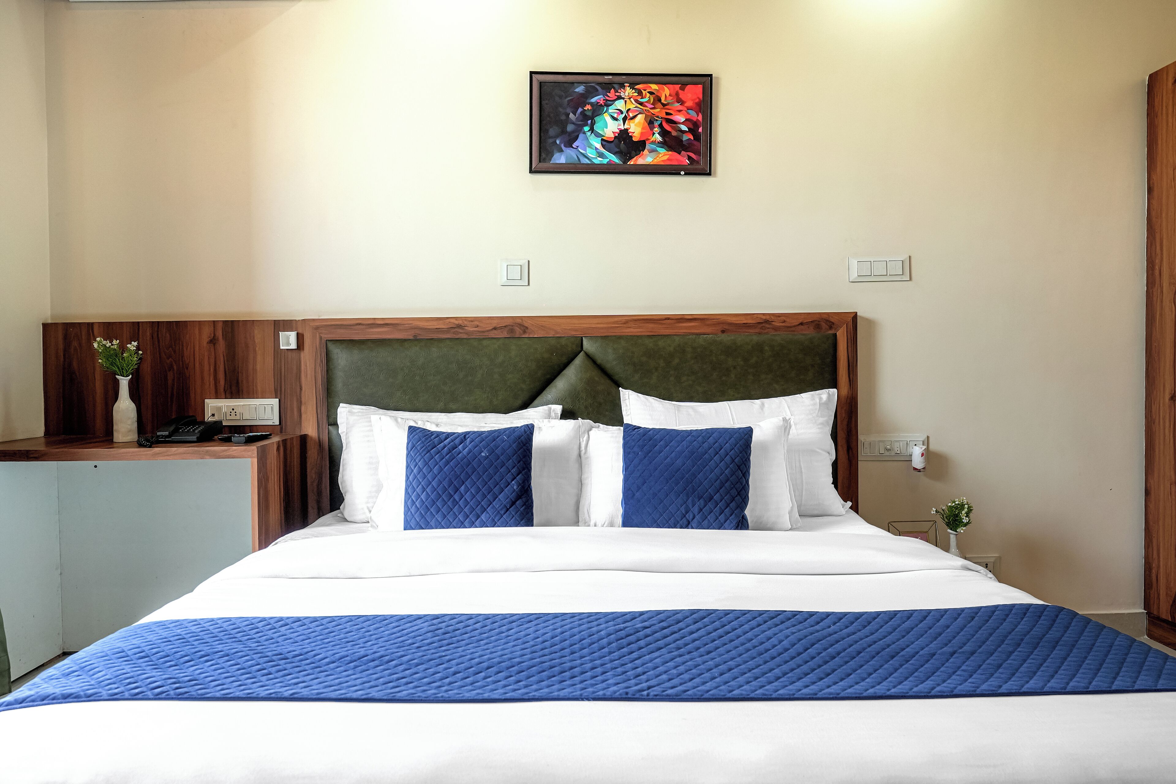 A hotel room with a bed, a wooden headboard, a bedside table with a vase and a phone, and a painting on the wall.