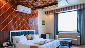 A modern hotel room with a wooden wall panel design, a large bed with white bedding, a ceiling fan, a window with curtains, and a seating area with a table. - Appas Hotel (Baijnath)