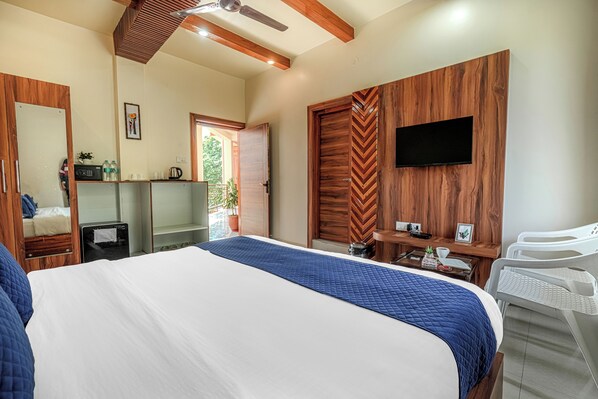 A bedroom with a large bed, a TV mounted on a wooden wall, a desk with a chair, and a small balcony visible through an open door.
