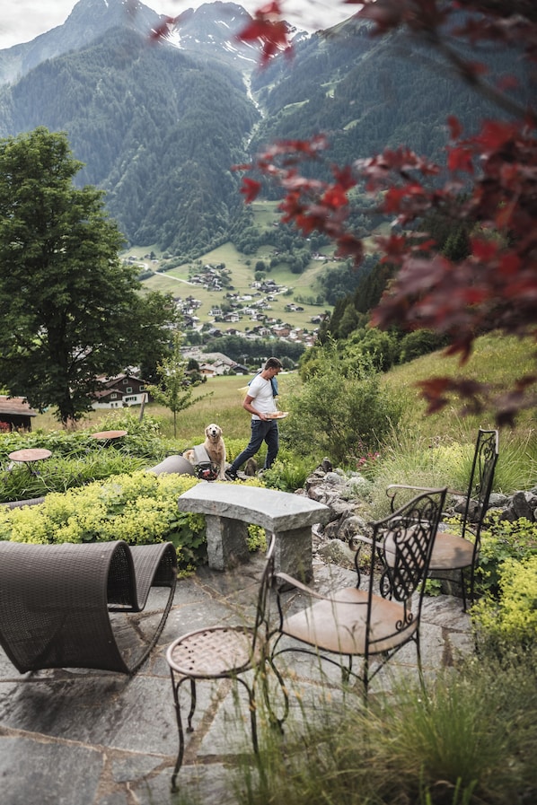 A person walking with a dog near outdoor seating with stone tables and chairs, surrounded by greenery and a mountainous backdrop.