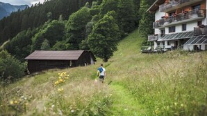 A person walking on a grassy path with a wooden cabin and a modern building with balconies and solar panels in the background. - So small and already 2 balconies, here the bedroom and living room with balcony. (Sankt Gallenkirch)