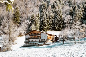 Un edifici d'alguns pisos amb façana de fusta, envoltat d'arbres coberts de neu i amb un paisatge de muntanyes al fons.