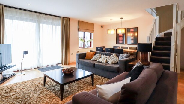 A modern living room with a sofa, coffee table, and a dining area in the background. - Lodge 22 Cherry (Wadebridge)