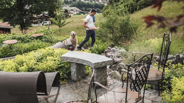 Eine Person geht mit einem Hund vorbei an Sitzgelegenheiten im Freien, mit Steintischen und -stühlen, umgeben von viel Grün und einer Bergkulisse.