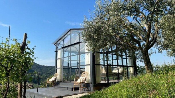 Ein modernes Haus aus Glas mit Terrasse und Sitzbereich, umgeben von Grünflächen und Hügeln.