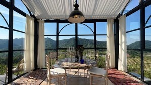 Ein Essbereich mit einem runden Tisch, Stühlen und einer Pendelleuchte. Große Fenster bieten Blick auf die Berge.