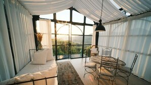 Ein gemütliches Zimmer mit einem Bett, einem Esstisch und Stühlen. Große Fenster bieten Blick ins Freie.