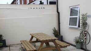 A wooden picnic table with benches in a courtyard with potted plants and a bicycle leaning against the wall. - The Old Bike Shop Accommodation (Norwich)