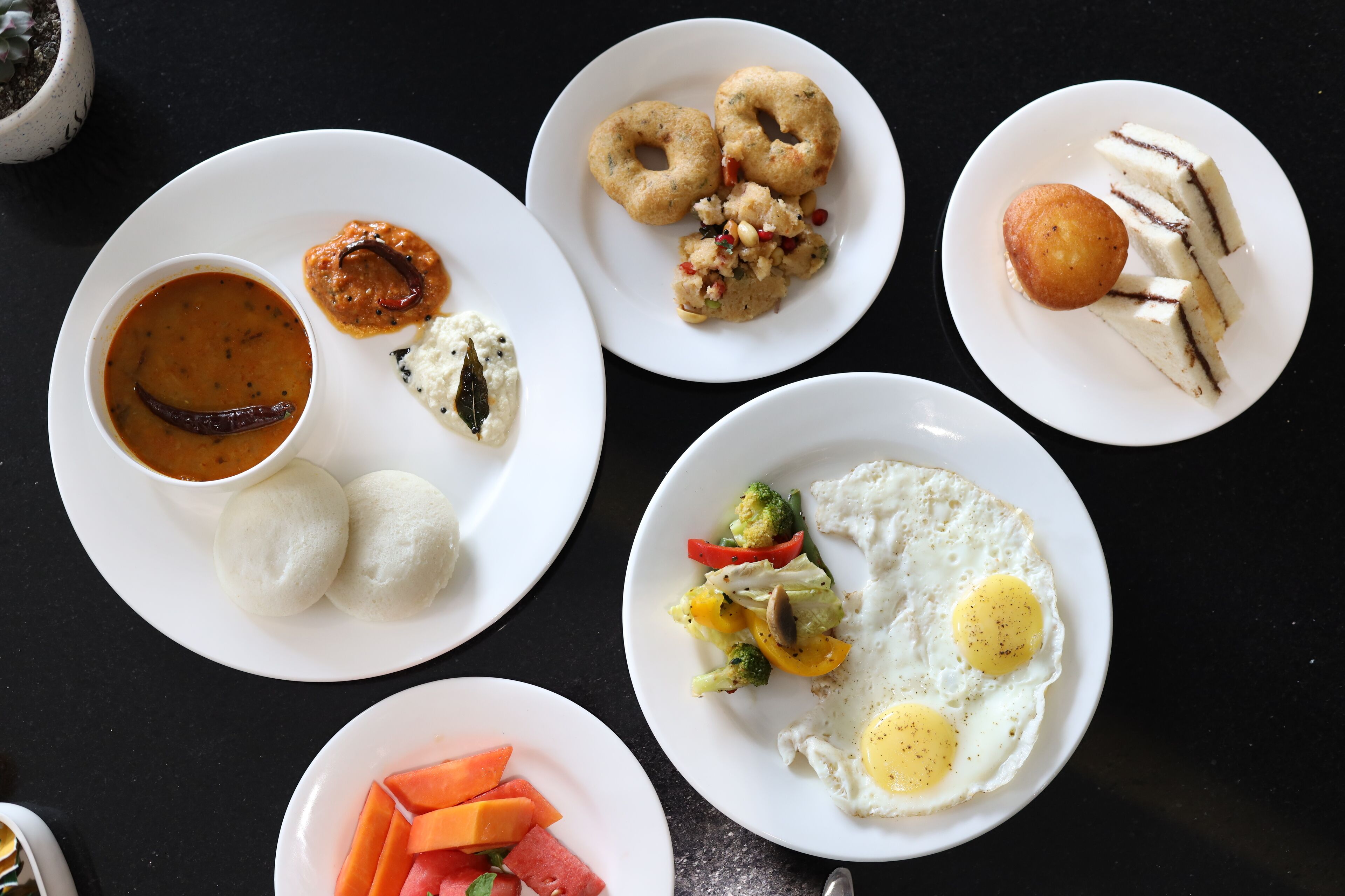 A variety of dishes on white plates, including idlis, sambar, vada, and a vegetable stir-fry with eggs.