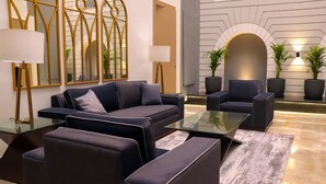 A modern living room with a dark-colored sofa, armchairs, and a glass coffee table. There are two floor lamps, a large mirror, and a fireplace with potted plants. - Ahuja Residency Mayfield Garden (Gurugram)