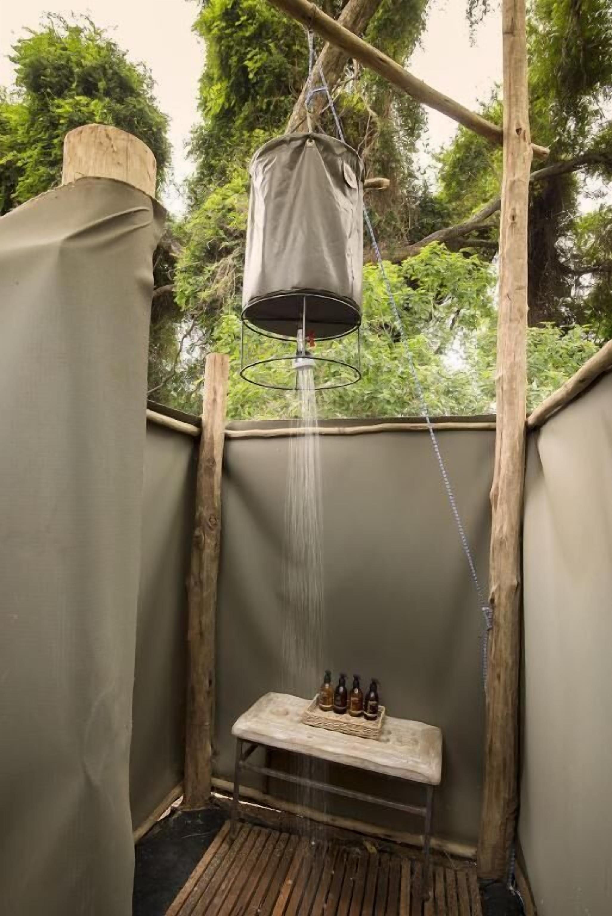 Une douche extérieure rustique, avec un récipient à eau suspendu, un banc en bois et des bouteilles posées sur le sol.