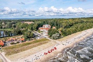 An aerial view of a coastal area with a beach, a large building with multiple red-roofed structures, and surrounding greenery. - Villa Cölpin Wohnung Stranddistel - Zentral und Strandnah Gelegen (Kölpinsee)