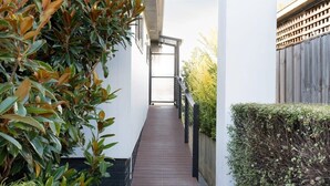 Uma passagem estreita que conduz à entrada de um edifício, ladeada por vegetação e um cercado de madeira.