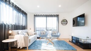 Uma sala de estar moderna com um sofá, duas poltronas, uma mesa de centro, uma televisão e uma janela grande com cortinas.