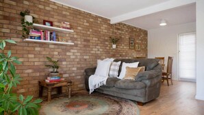 Un salón con pared de ladrillo, un sofá gris, un tapete estampado y una estantería con libros y objetos decorativos.
