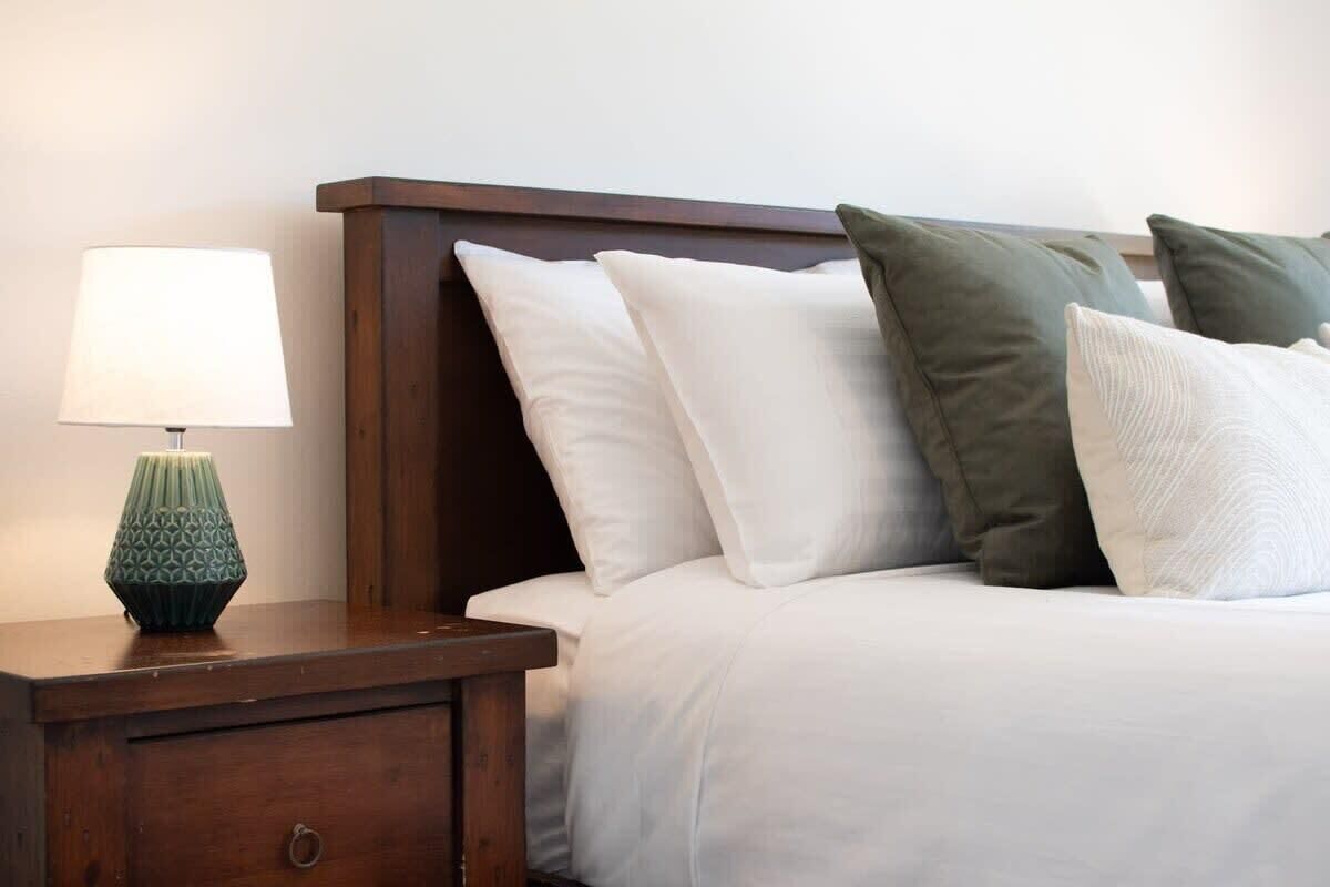 Een net opgemaakt bed met witte beddengoed en een houten hoofdbord. Er staat een nachtkastje met een lamp en een decoratief object.